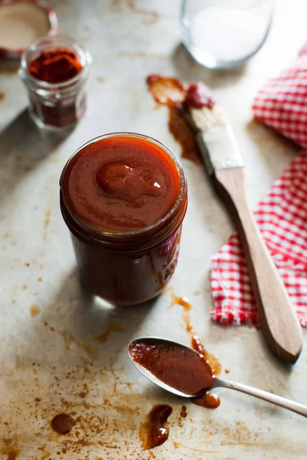 Paleo BBQ sauce in a mason jar on a metal table with a spoon in front with sauce in it, a basting brush and a red checkered napkin