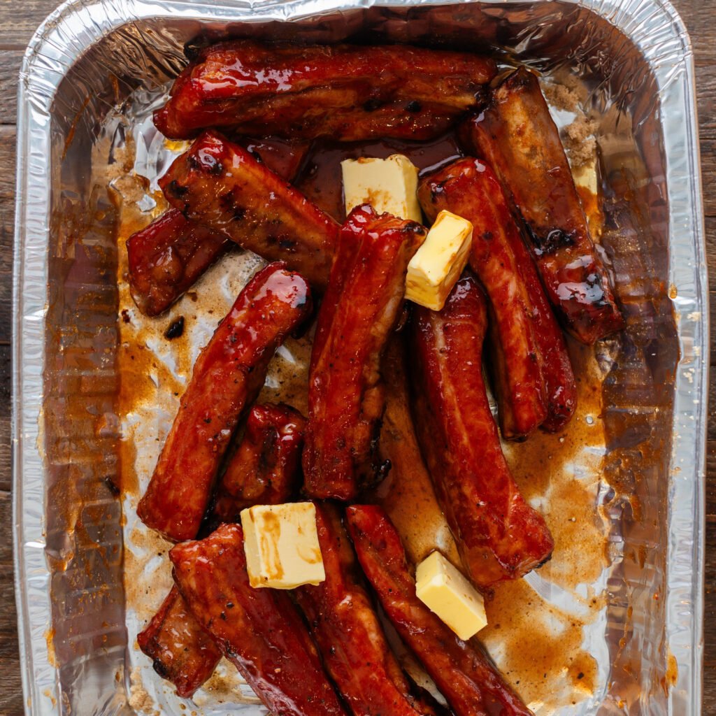 ribs in aluminum pan being tossed with bbq sauce, butter, honey and brown sugar