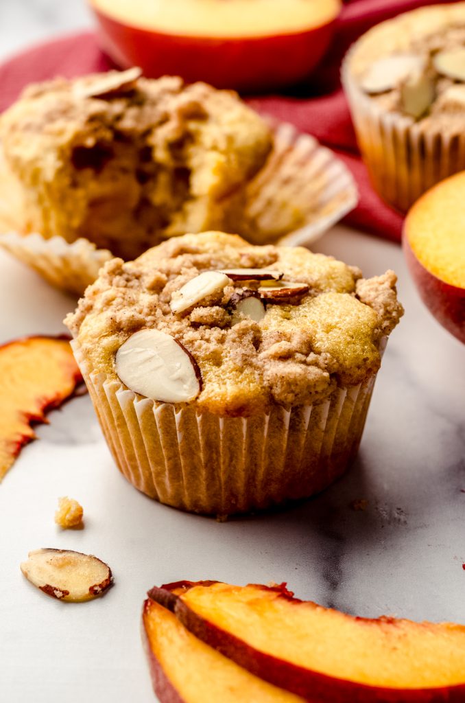 A peach almond streusel muffin sitting on a surface with peaches and almond slices around it.