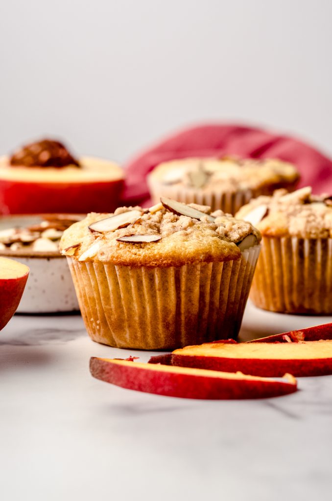 A peach almond streusel muffin sitting on a surface with peaches and almond slices around it.
