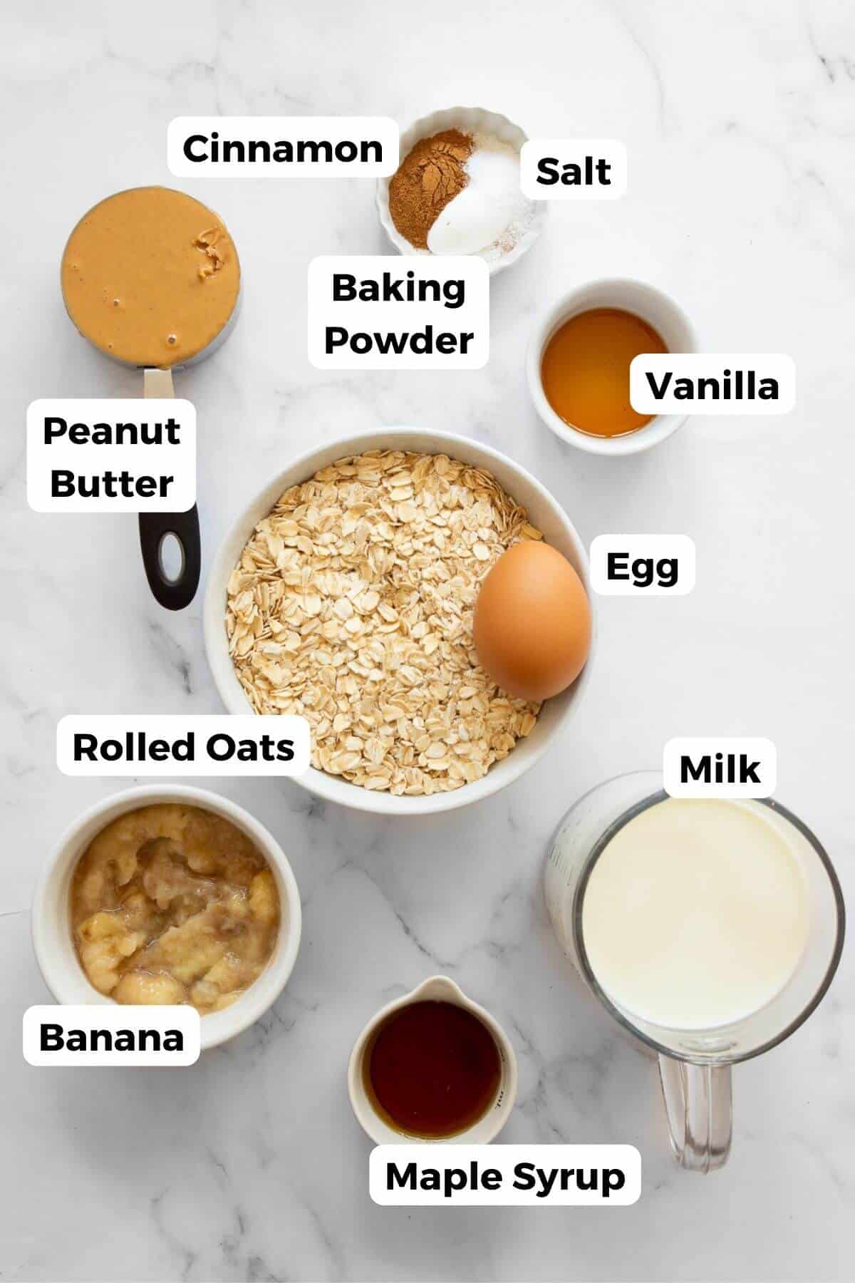 All the ingredients for peanut butter banana baked oatmeal arranged in small bowls with labels, including oats, peanut butter, banana, eggs, milk, maple syrup, cinnamon, and baking powder.