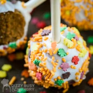 Side close up shot of a pumpkin cake pop with fall spinkles on a black surface with more cake pops in the background.