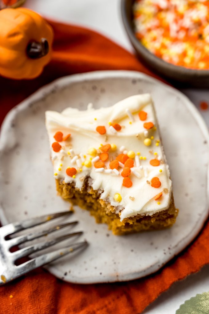 A slice of pumpkin sheet cake sitting on a plate with a fork and the cake has a bite taken out of it.