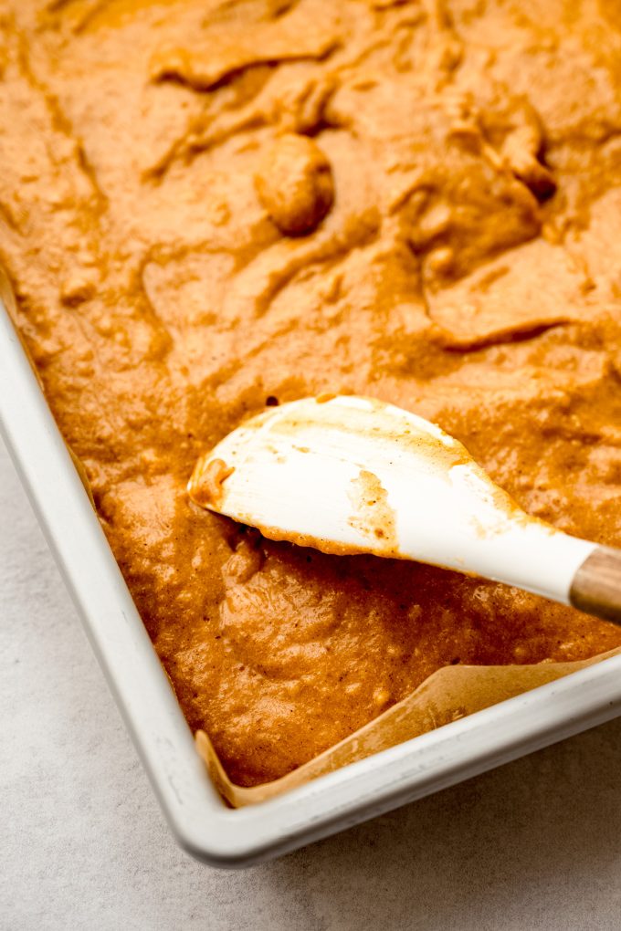Someone is using a spatula to spread pumpkin cake batter into a sheet cake pan.