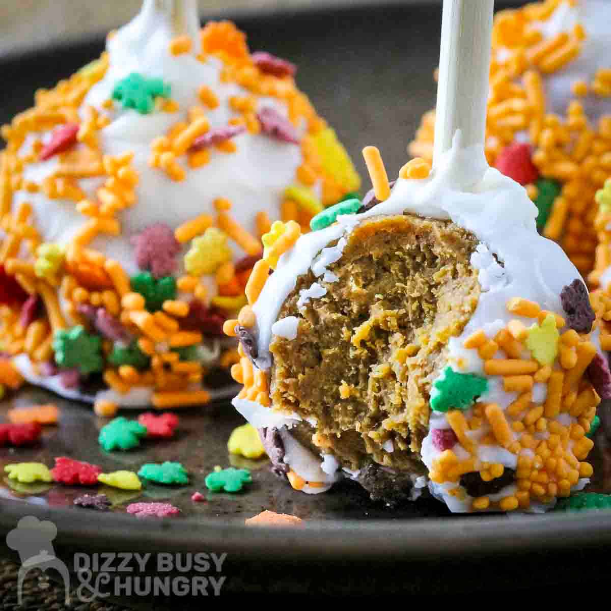 Side shot of a pumpkin cake pop with fall sprinkles and a bite taken out on a black plate with more cake pops in the background.