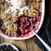 Apple blackberry crumble being scooped out of a white ceramic dish with a scoop of vanilla ice cream on top.