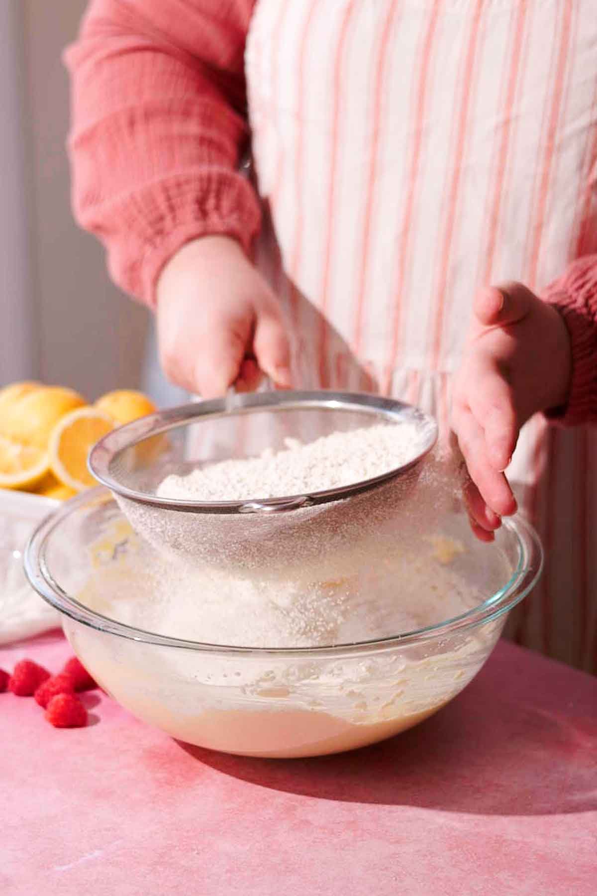 sifting flour with a metal sieve into a glass bowl of batter