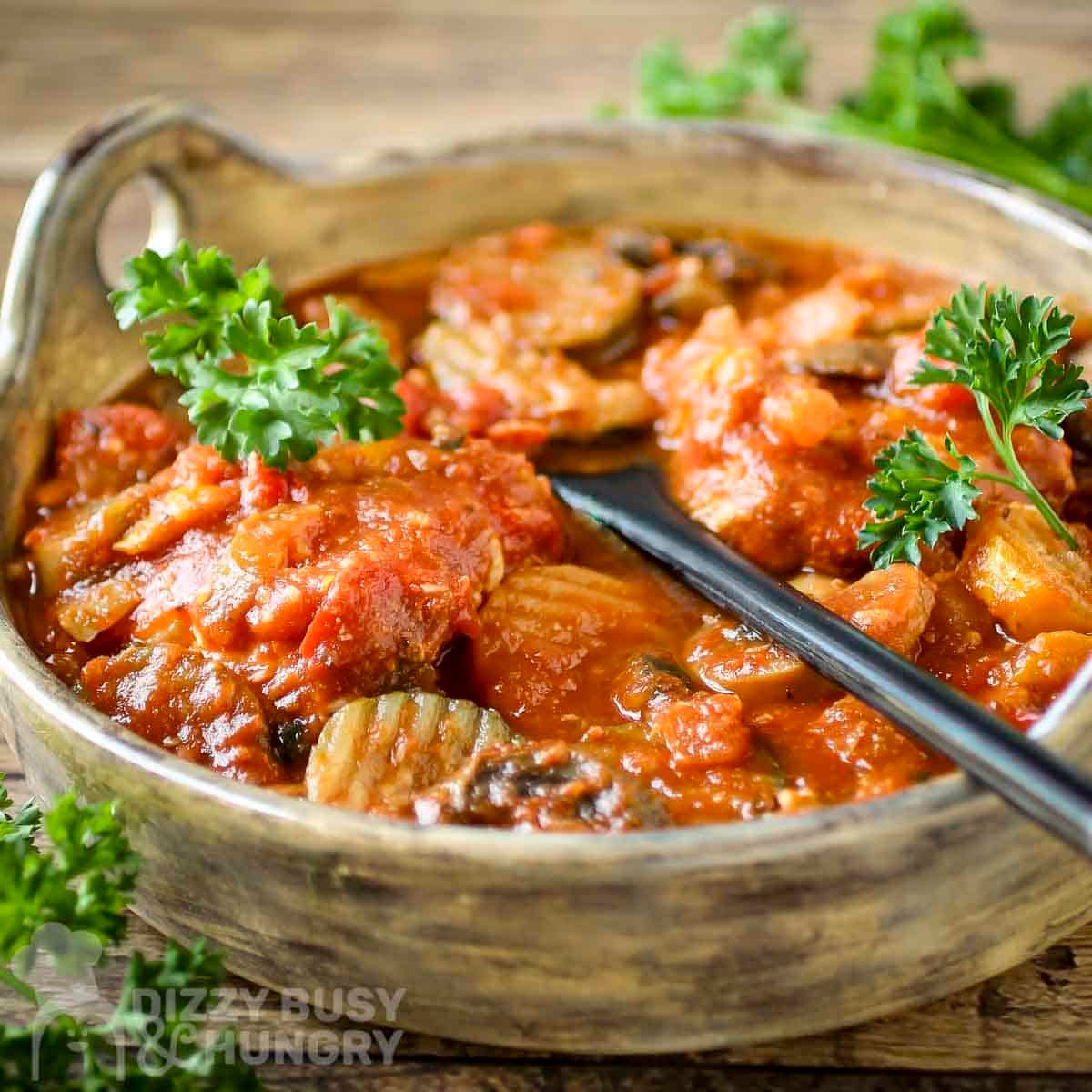 Side close up shot of chicken cacciatore in a large serving bowl with a black spoon inside on a wooden surface with a bunch of fresh parsley on the side.