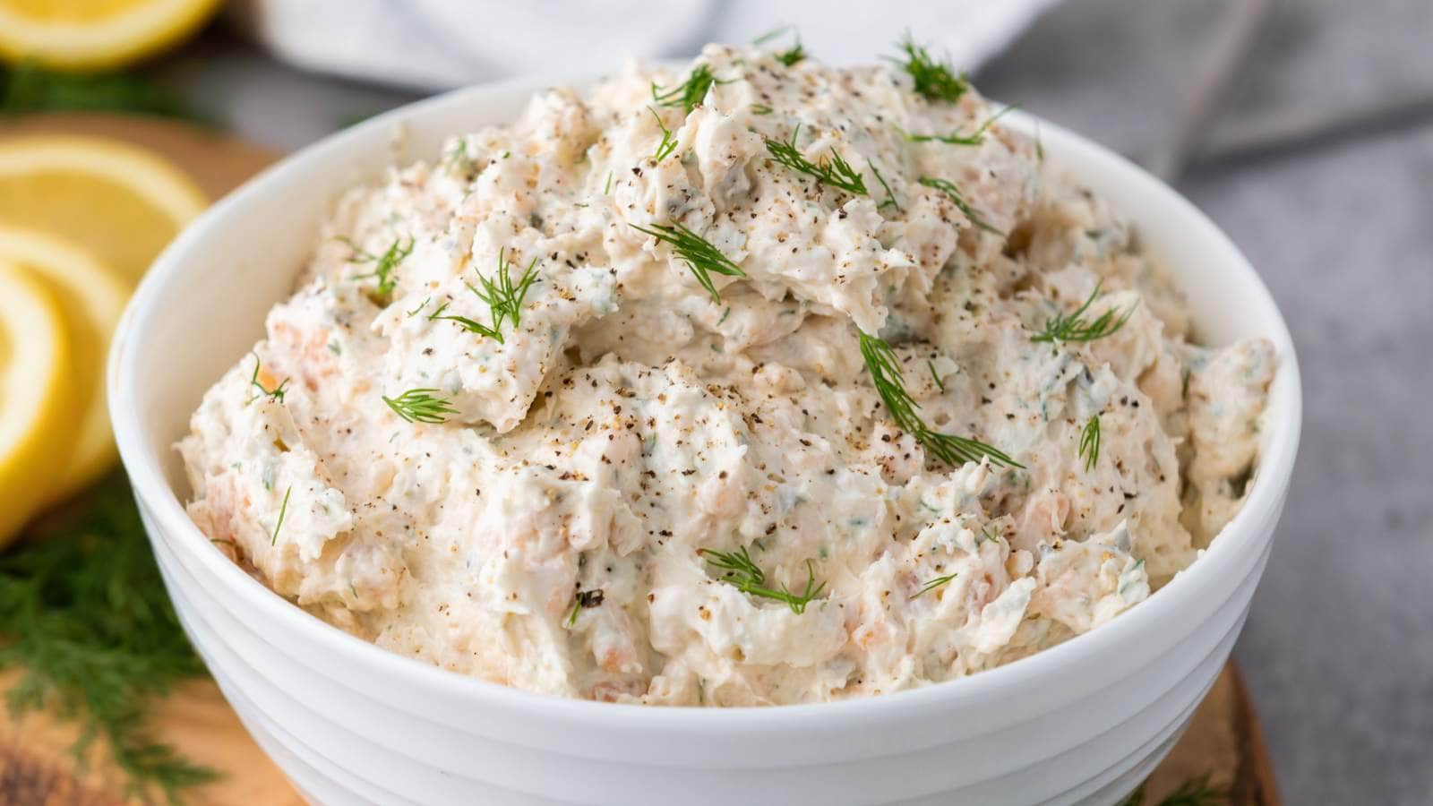 A white bowl filled with salmon dip with cream cheese, garnished with dill and ground pepper.