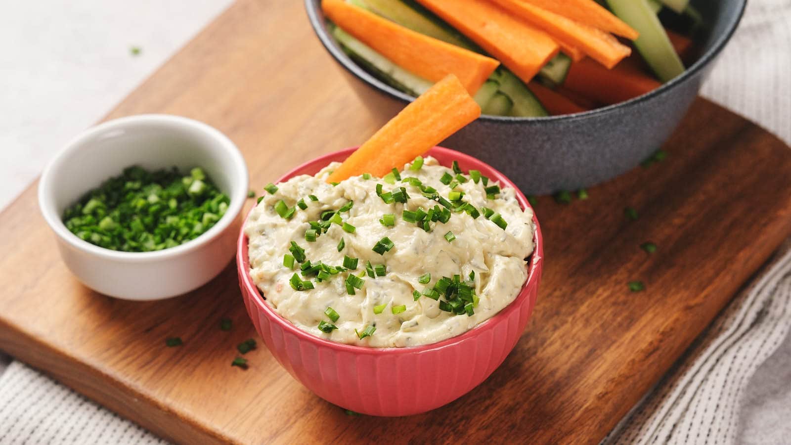 A bowl of sour cream dip garnished with chopped chives, accompanied by carrot and cucumber sticks in a bowl.