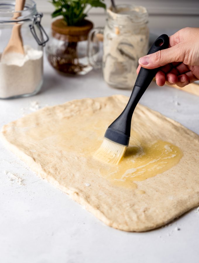 Someone is brushing melted butter onto a rectangle of sourdough cinnamon roll dough.