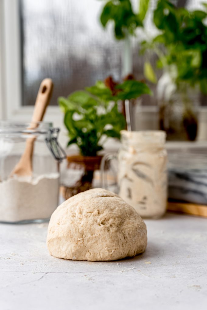A ball of sourdough starter cinnamon roll dough on a table with a canister of flour and jar of starter in the background.