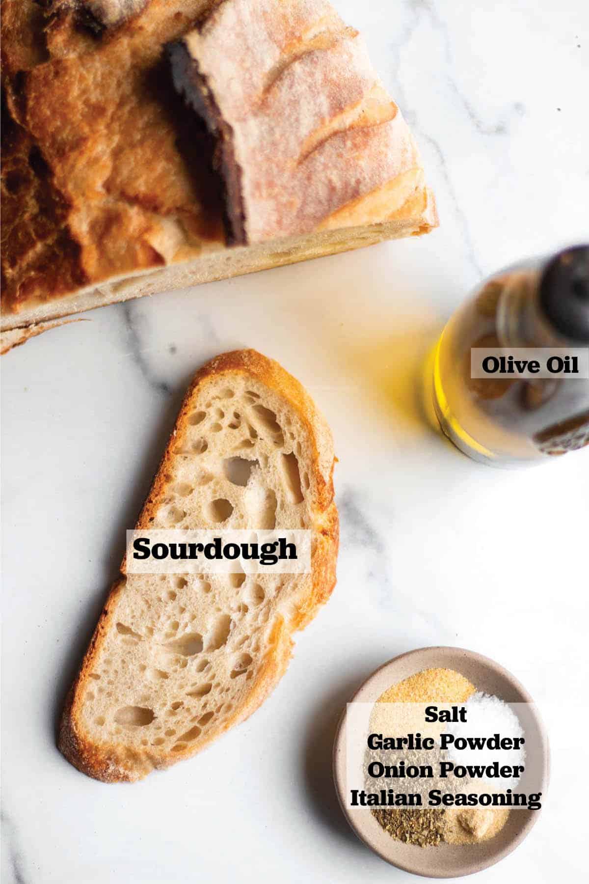 Half a loaf of sourdough bread with a single slice, bottle of olive oil and a dish with salt, Italian seasoning, garlic powder and onion powder. 
