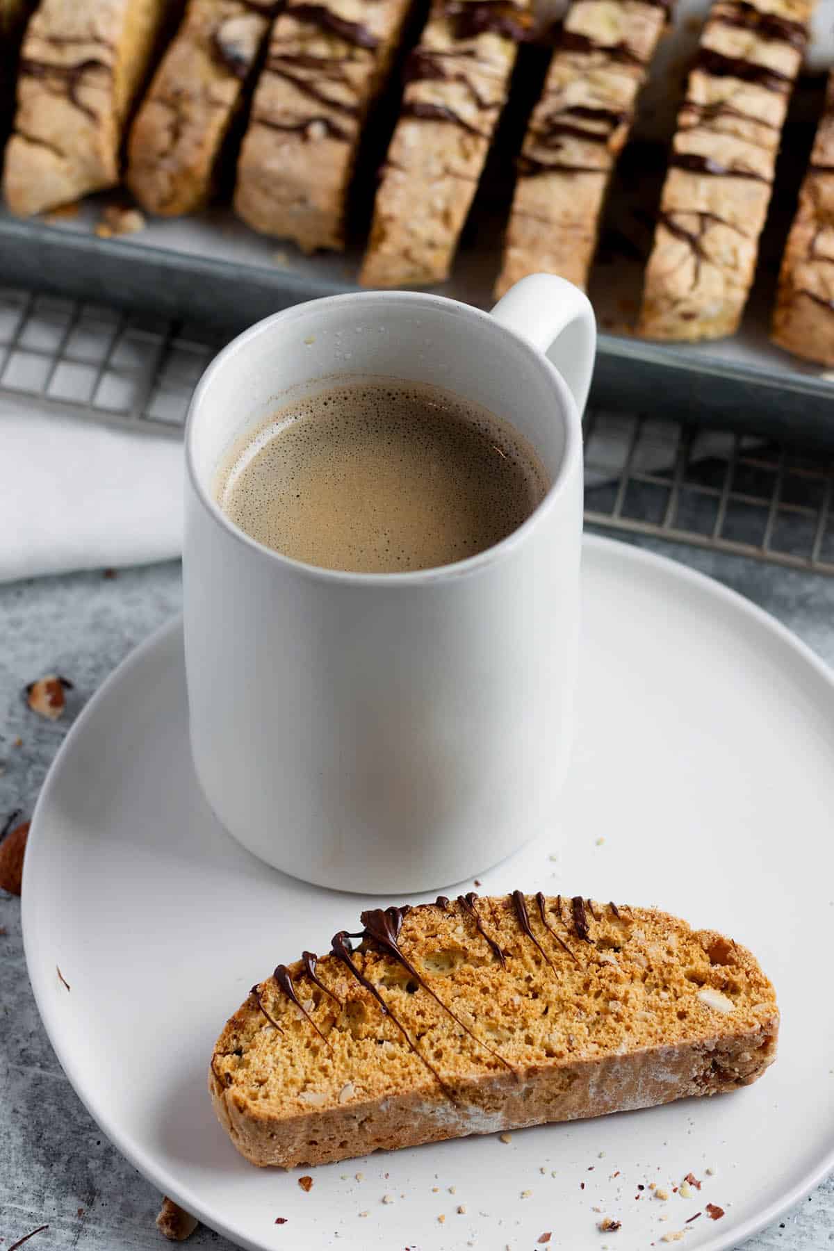 A plate with a piece of almond biscotti, a mug of coffee and a metal rack with more biscotti in the back. 