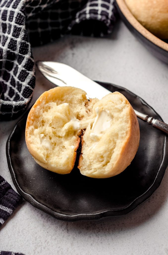 A sourdough dinner roll sitting on a black plate. It has been split apart and spread with a little butter. There is a butter knife behind it.