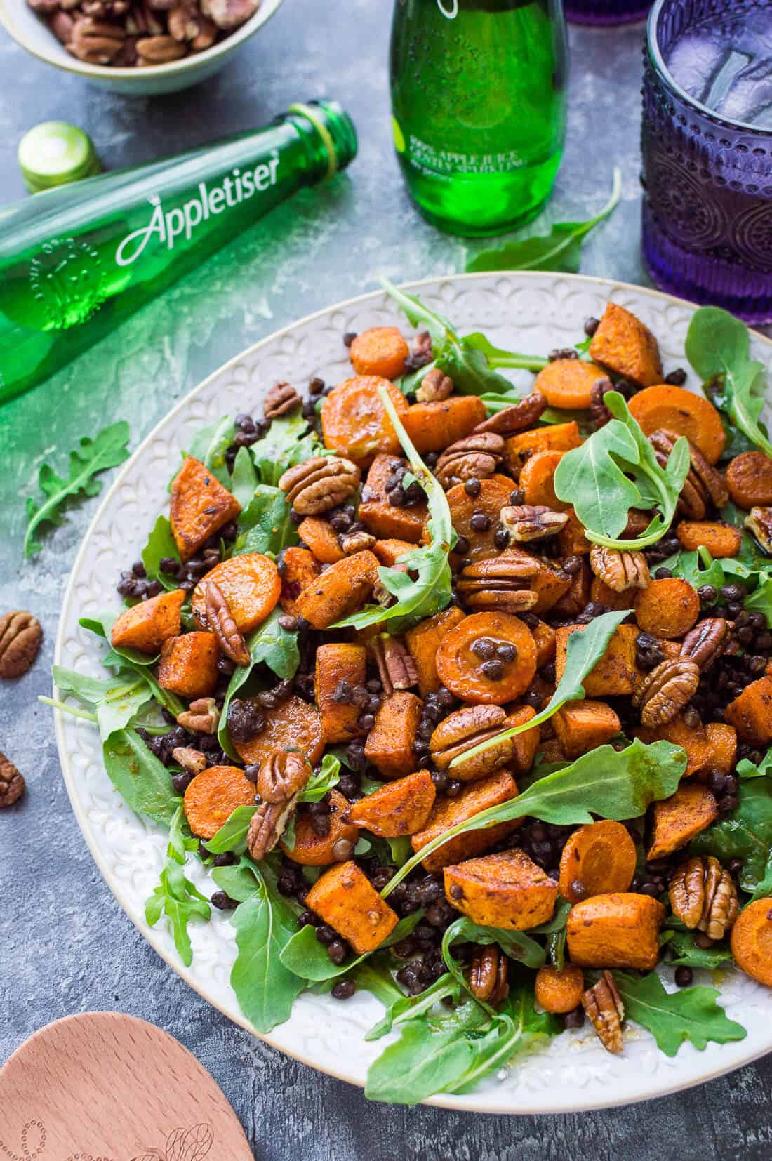 Roasted sweet potato, lentil and rocket salad with toasted pecans on a white plate with Appletiser on a grey background.