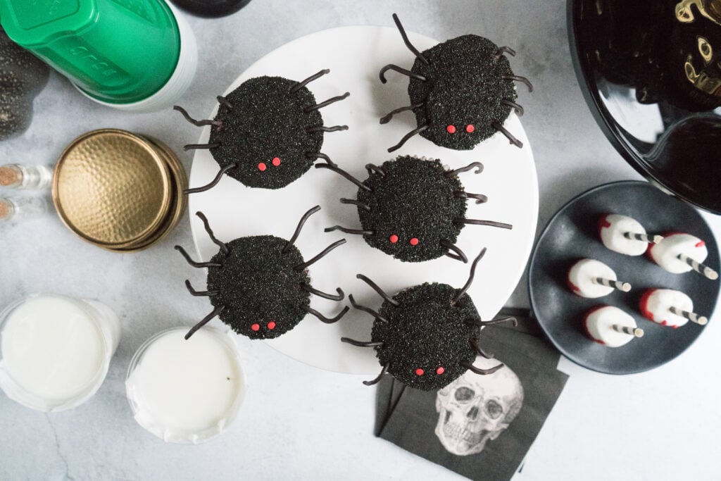 Top-down view of festive Halloween spider cupcakes on a white cake stand. Each cupcake is coated in black sprinkles with two small red candy eyes and eight chocolate licorice legs. The spooky display is surrounded by glasses of milk, black skull napkins, and other Halloween-themed treats on a light gray surface.