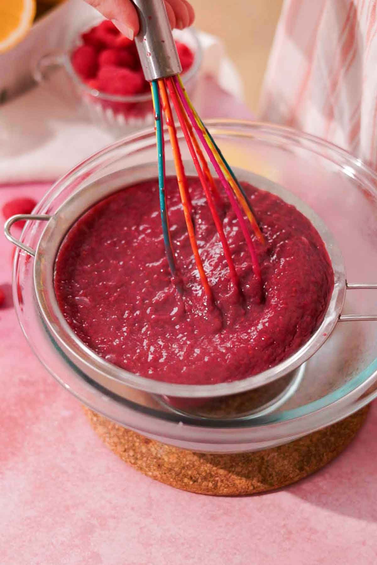 straining raspberry curd with silicone whisk through a metal sieve on top of a glass bowl