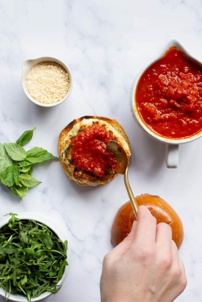 adding warmed marinara sauce on top of cooked chicken parmesan burgers