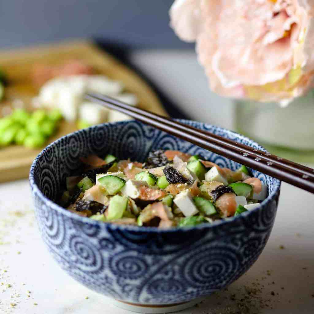 Photo of white marble counter with blue decorated bowl filled with sushi rice and vegan toppings (tofu, cucumber, seaweed, sushi ginger, etc) and flowers and ingredients on a chopping board in the background.