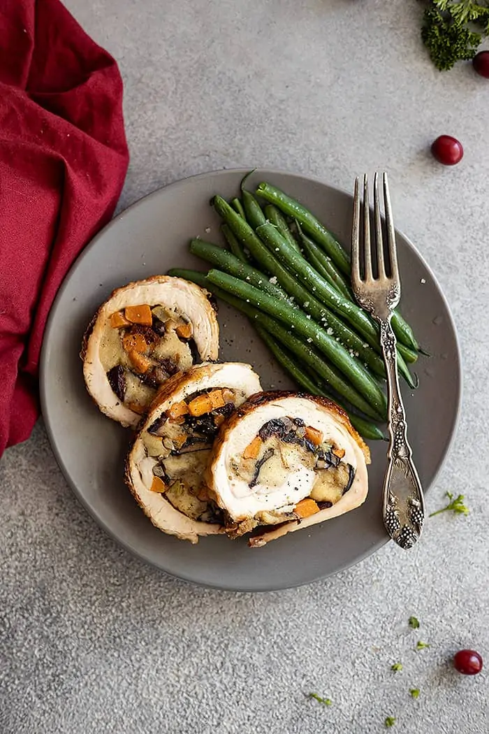 Overhead view of three slices of sweet potato and stuffing turkey roulade on a plate with a side of green beans. A red napkin is off to the side.