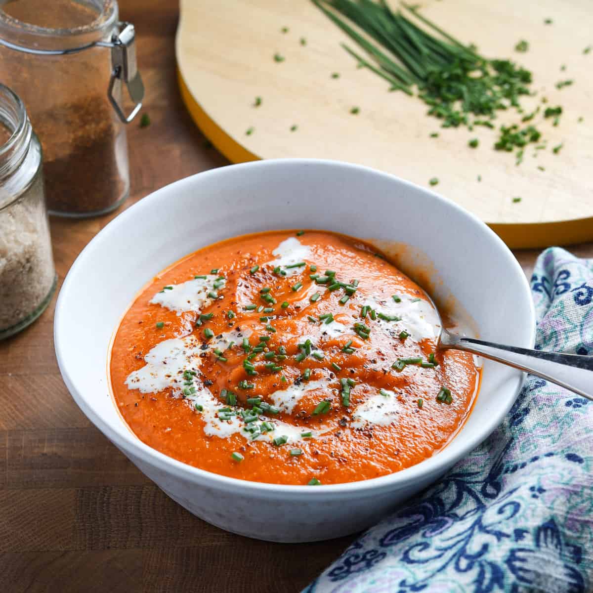 Photo of a bowl of tomato, lentil and squash soup on a wooden table with a blue napkin, jars of cayenne and smoked salt and a chopping board with chives in the background.