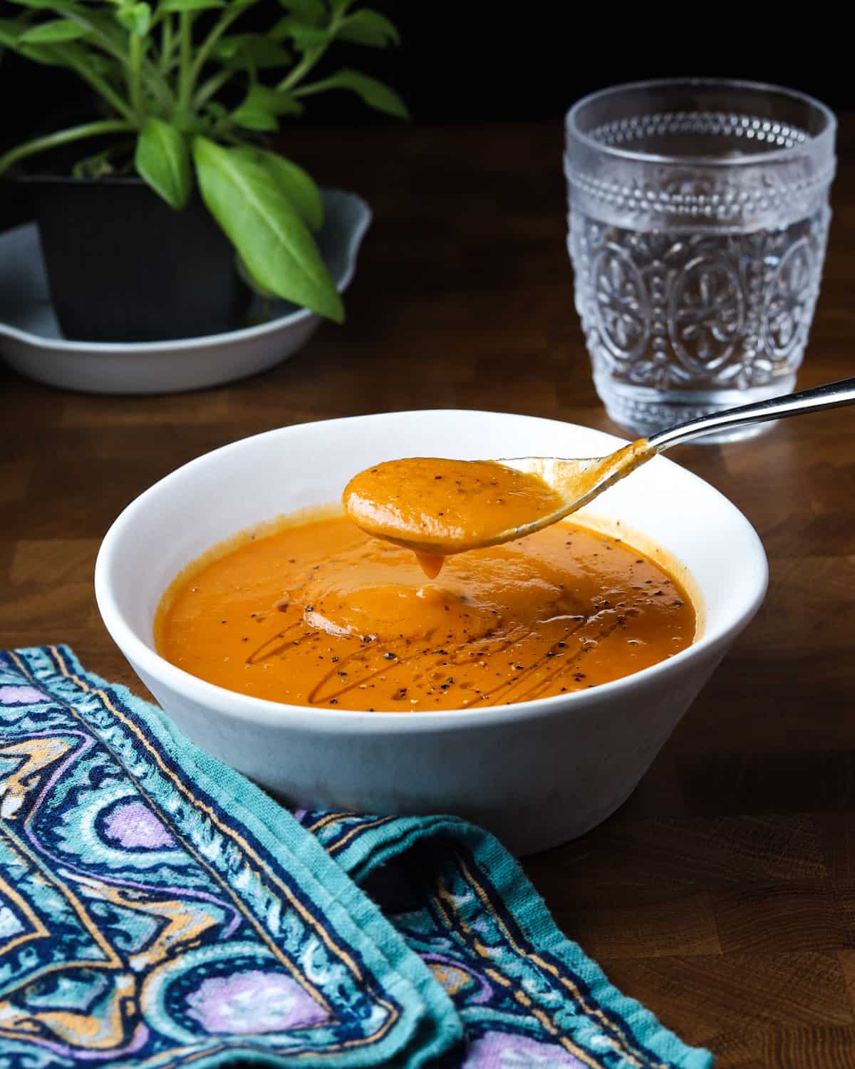 Photo of someone taking a spoonful of tomato and lentil soup form a white bowl on a wooden table with teal patterned napkin, glass of water and plant in the background.
