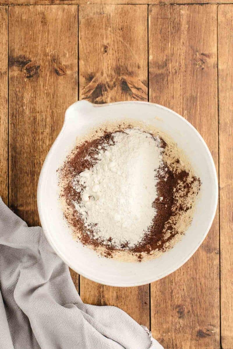 A white mixing bowl containing flour and cocoa powder sits on a wooden surface next to a light gray cloth.