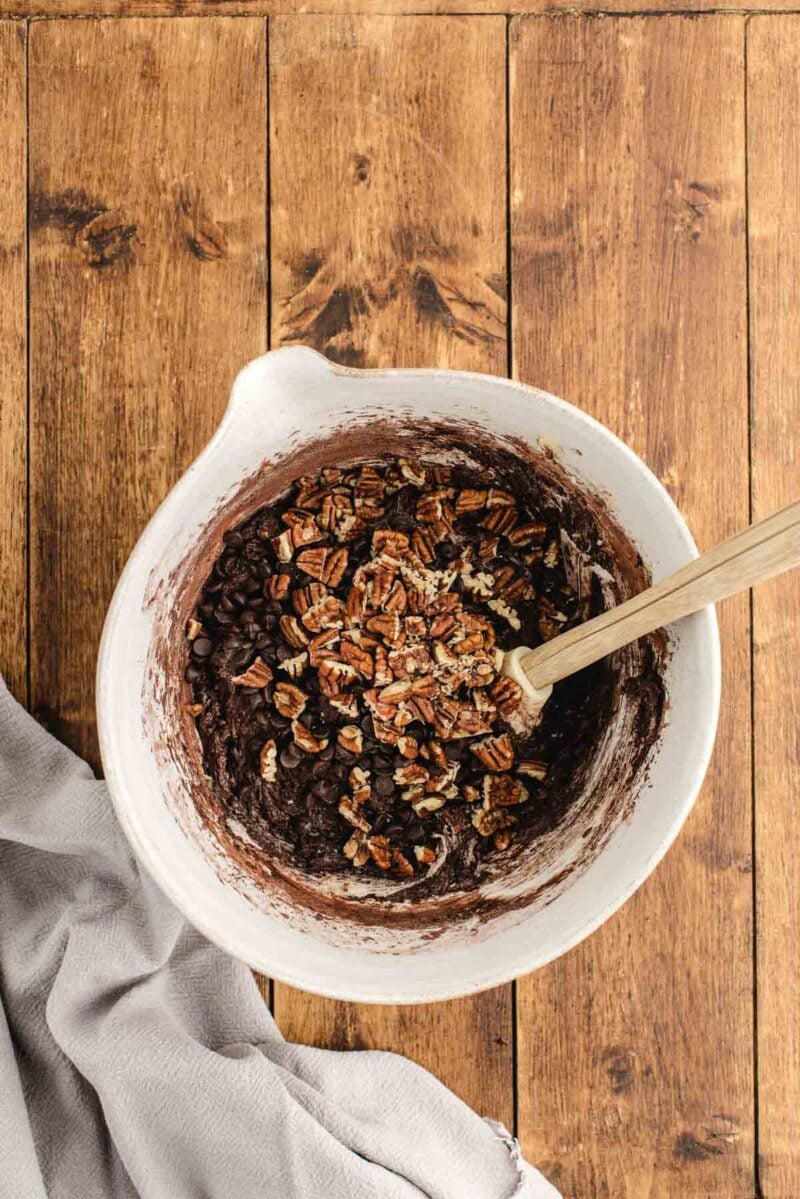 A mixing bowl filled with chocolate batter, chocolate chips, and chopped pecans on a wooden surface, with a wooden spoon and a gray cloth nearby.