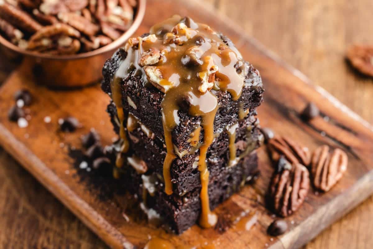 Three stacked chocolate turtle brownies topped with chopped pecans and drizzled with caramel sauce on a wooden board, with extra pecans and chocolate chips scattered around.