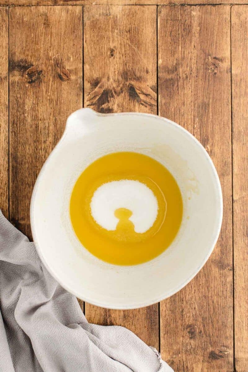 A white mixing bowl with melted butter and sugar sits on a wooden surface next to a light gray cloth.