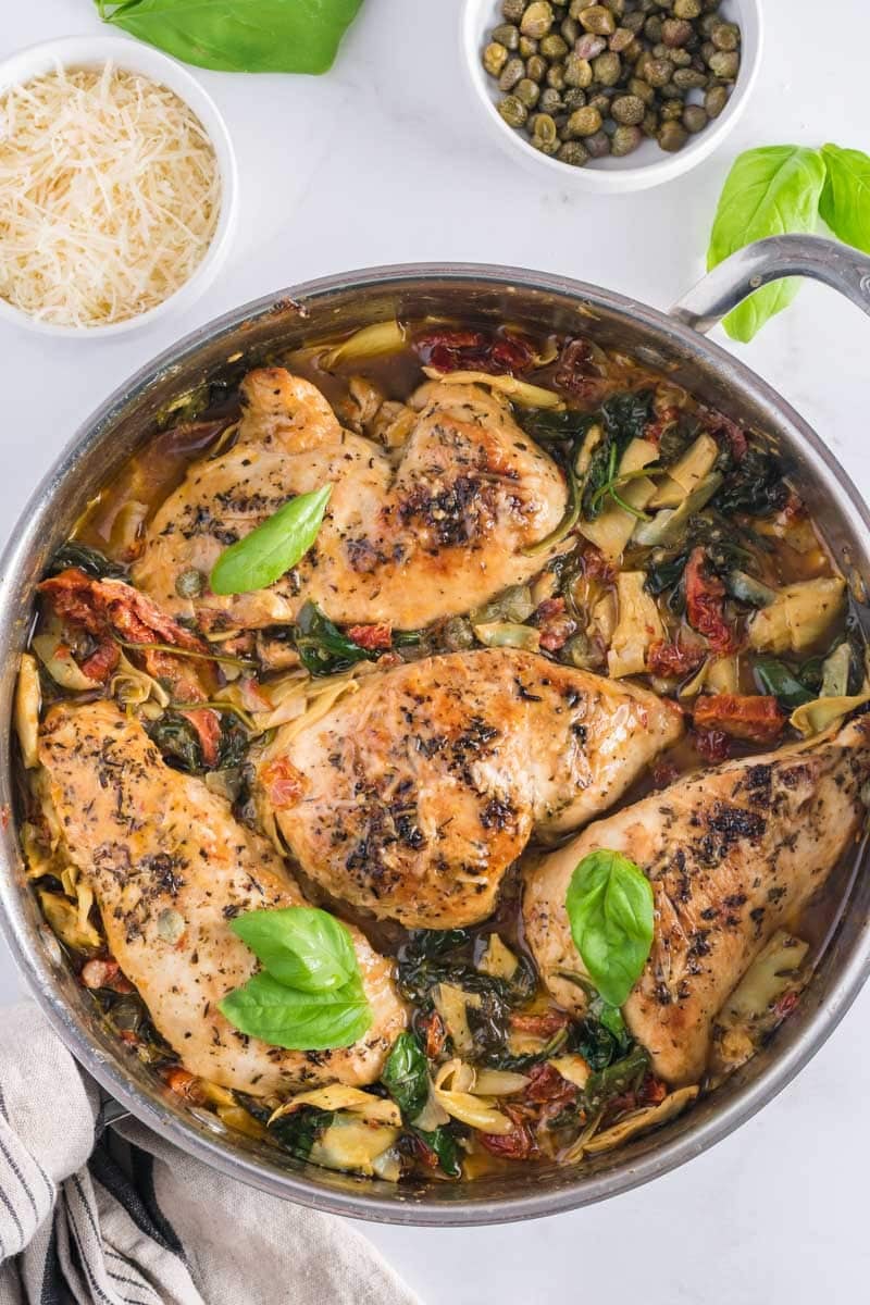 A skillet filled with cooked chicken breasts, spinach, sun-dried tomatoes, and artichoke hearts, garnished with fresh basil leaves. Parmesan and capers are in small bowls nearby.