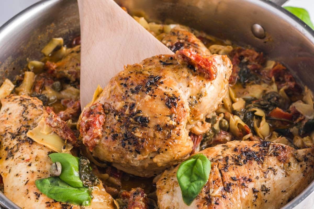 Cooked seasoned chicken breasts in a skillet with vegetables, herbs, sun-dried tomatoes, and a wooden spatula.