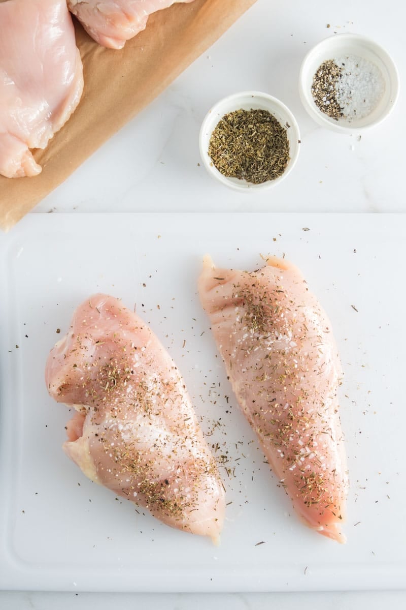 Two raw chicken breasts on a white cutting board, sprinkled with herbs and seasoning; more raw chicken and small bowls of herbs and salt are nearby.