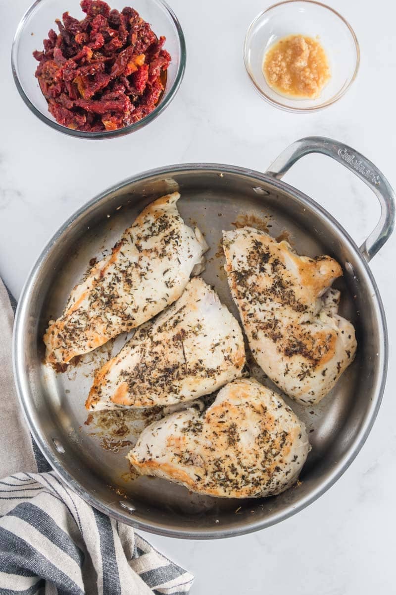 Four seasoned chicken breasts cooking in a stainless steel pan, with bowls of sun-dried tomatoes and minced garlic nearby on a white countertop.