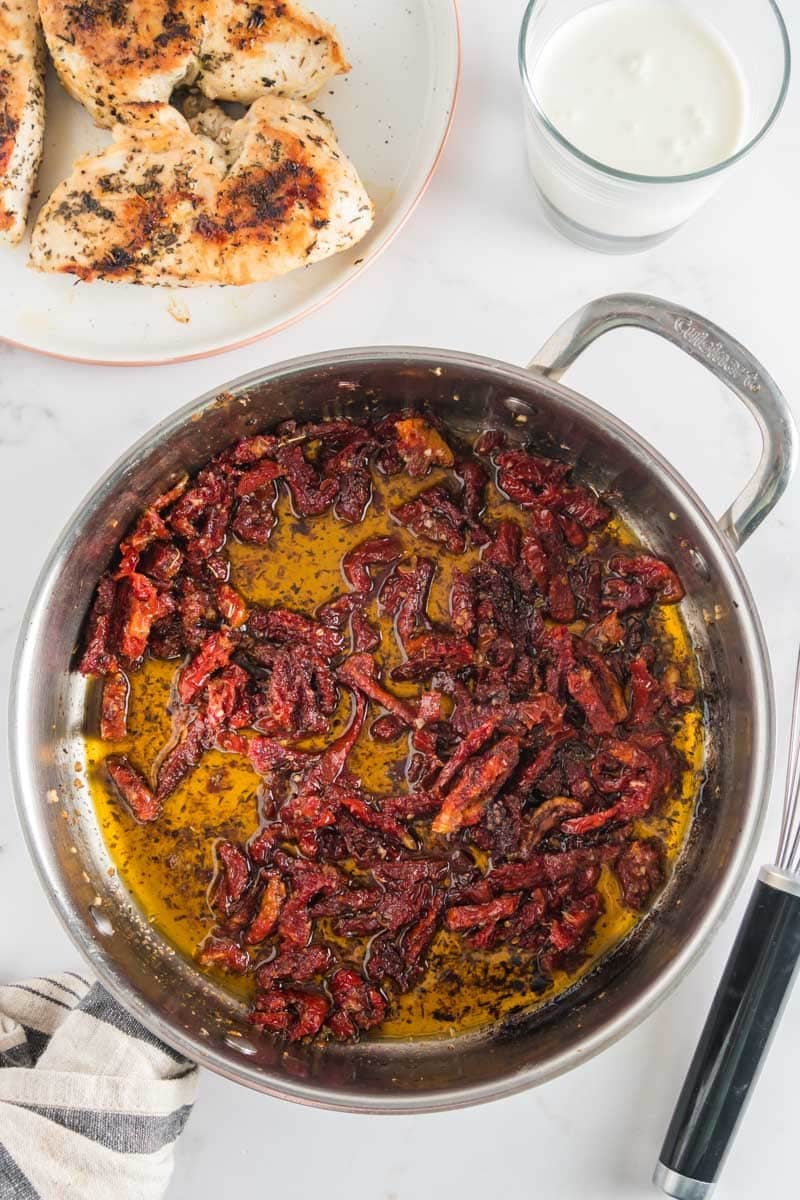A skillet filled with sun-dried tomatoes in oil sits on a white surface next to a plate of cooked chicken breasts and a glass of milk.