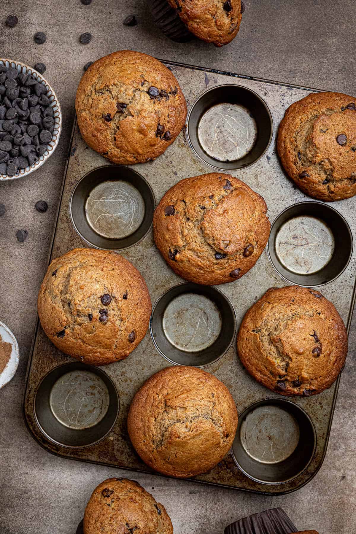 Vegan banana muffins in a muffin pan with a bowl of chocolate chips.