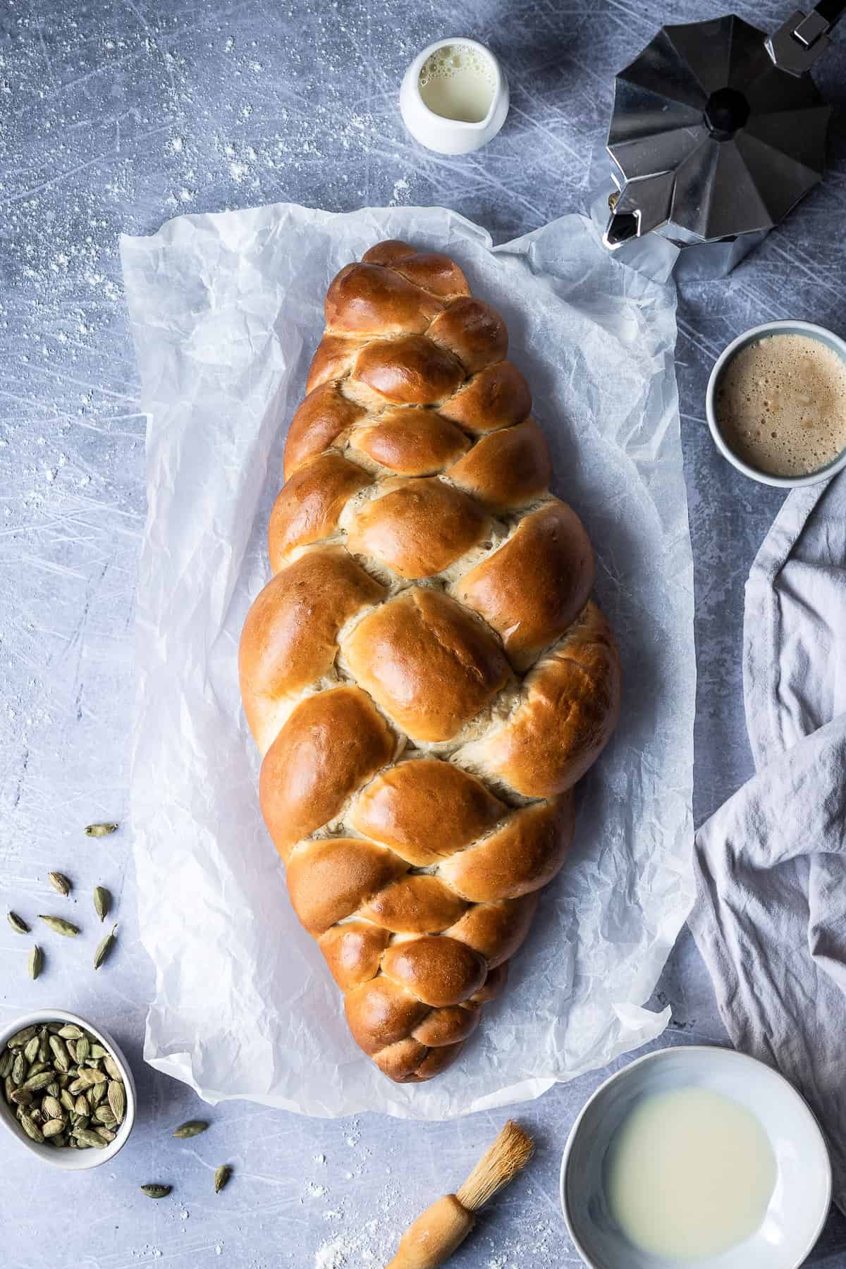The loaf of vegan pulla bread on a sheet of white baking parchment on a metal surface surrounded by a bowl of cardamom pods, bowl of milk, pastry brush, grey cloth, cup of coffee, caffetiere and jug of milk.
