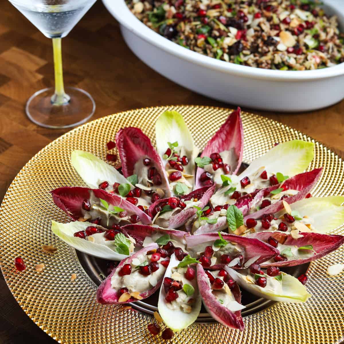Photo of vegan chicory canapés set out on a gold plate on a wooden tabletop with a cocktail and dish of wild rice salad in the background.