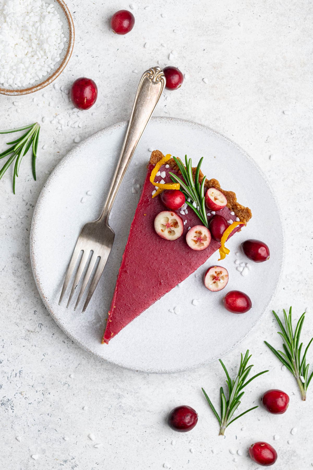 A slice of vegan cranberry curd tart on a white plate with fresh cranberries and sprigs of rosemary.