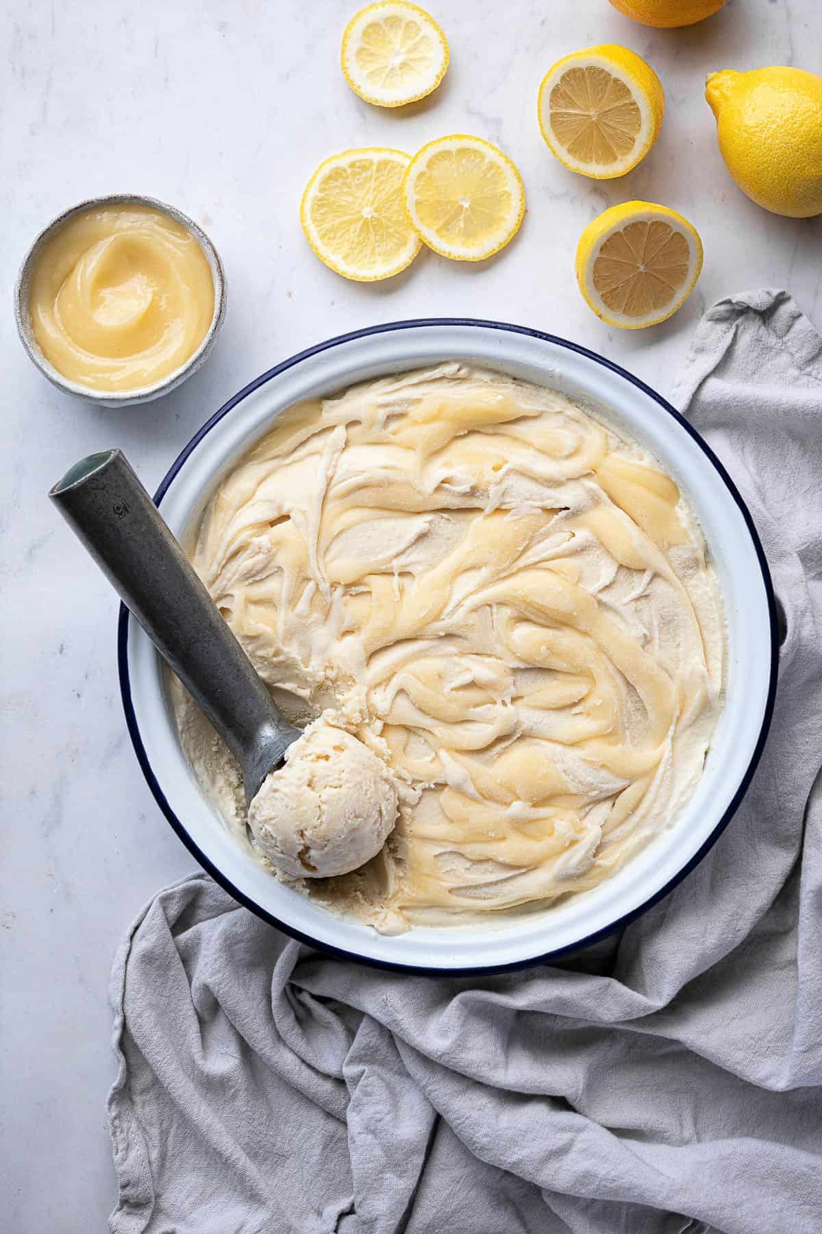 Vegan lemon ice cream in a bowl with an ice cream scoop, sliced lemons and bowl of lemon curd.