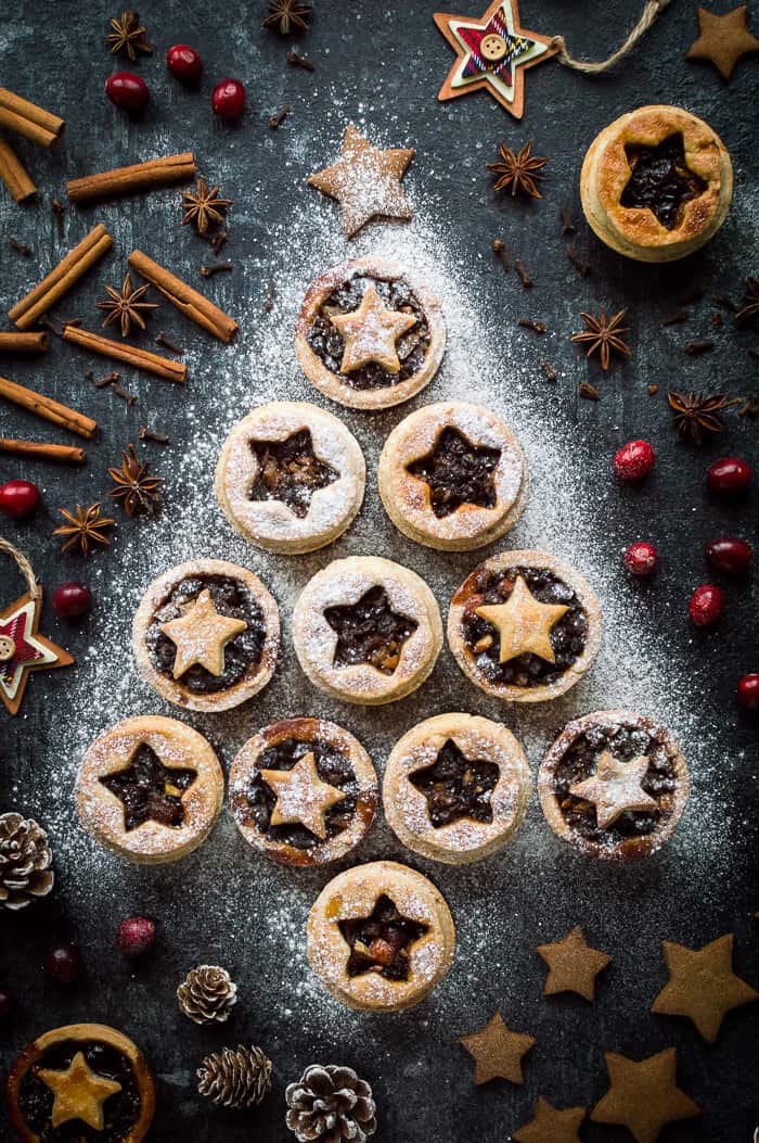 Vegan mince pies with coconut oil pastry arranged in a Christmas tree shape and sprinkled with icing sugar on a dark background with festive decorations.