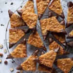 Vegan nussecken on a round wire cooling rack with a bowl of chocolate and some scattered hazelnuts.