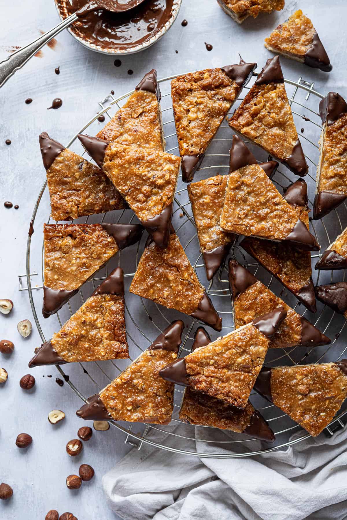 Vegan nussecken on a round wire cooling rack with a bowl of chocolate and some scattered hazelnuts.