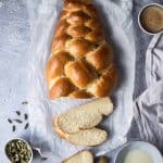 Vegan pulla bread with three pieces sliced off on a sheet of baking parchment on a metal surface with a bowl of cardamom pods, bowl of milk and pastry brush, cup of coffee and a caffetiere.
