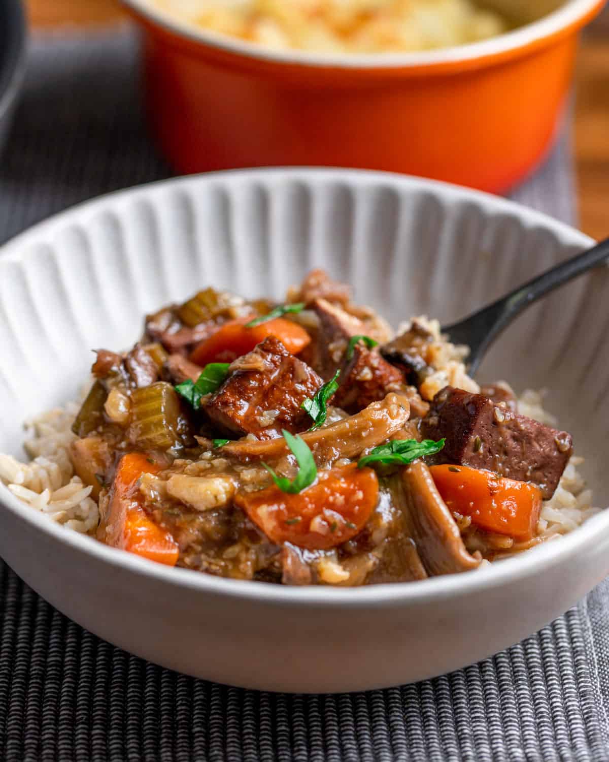 Photo of a bowl of vegan stew served with brown rice and a spoon, set on a grey mat with an orange pot in the background.