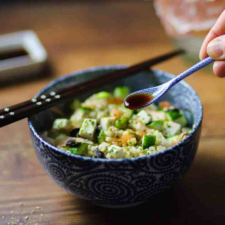 photo of a bowl of brown sushi rice with vegan sushi toppings (tofu, cucumber, sushi ginger slices, seaweed, etc.) and a spoonful of tamari sprinkling on top - with chopsticks resting on top.