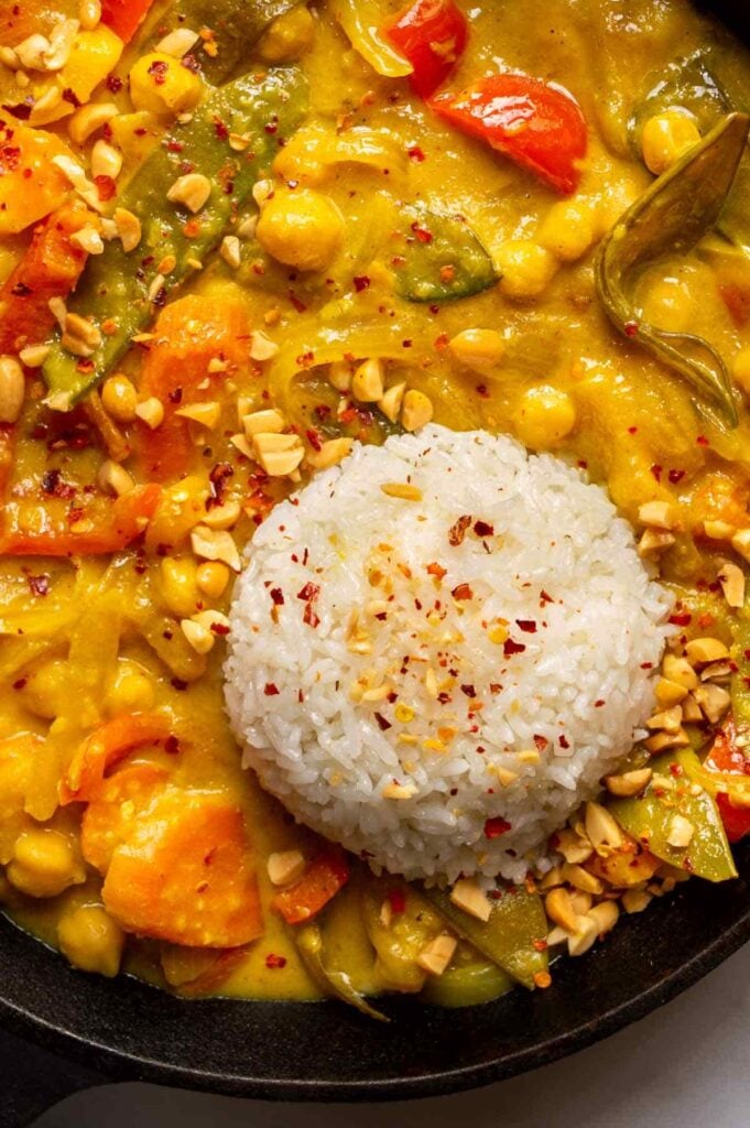 Close-up of vegetables in a yellow creamy curry sauce in a pan, with a pat of white rice on top.