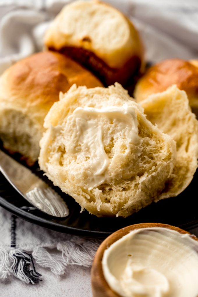 A yeast roll that has been split in half and buttered sitting on a plate with butter in the foreground and more rolls in the background.